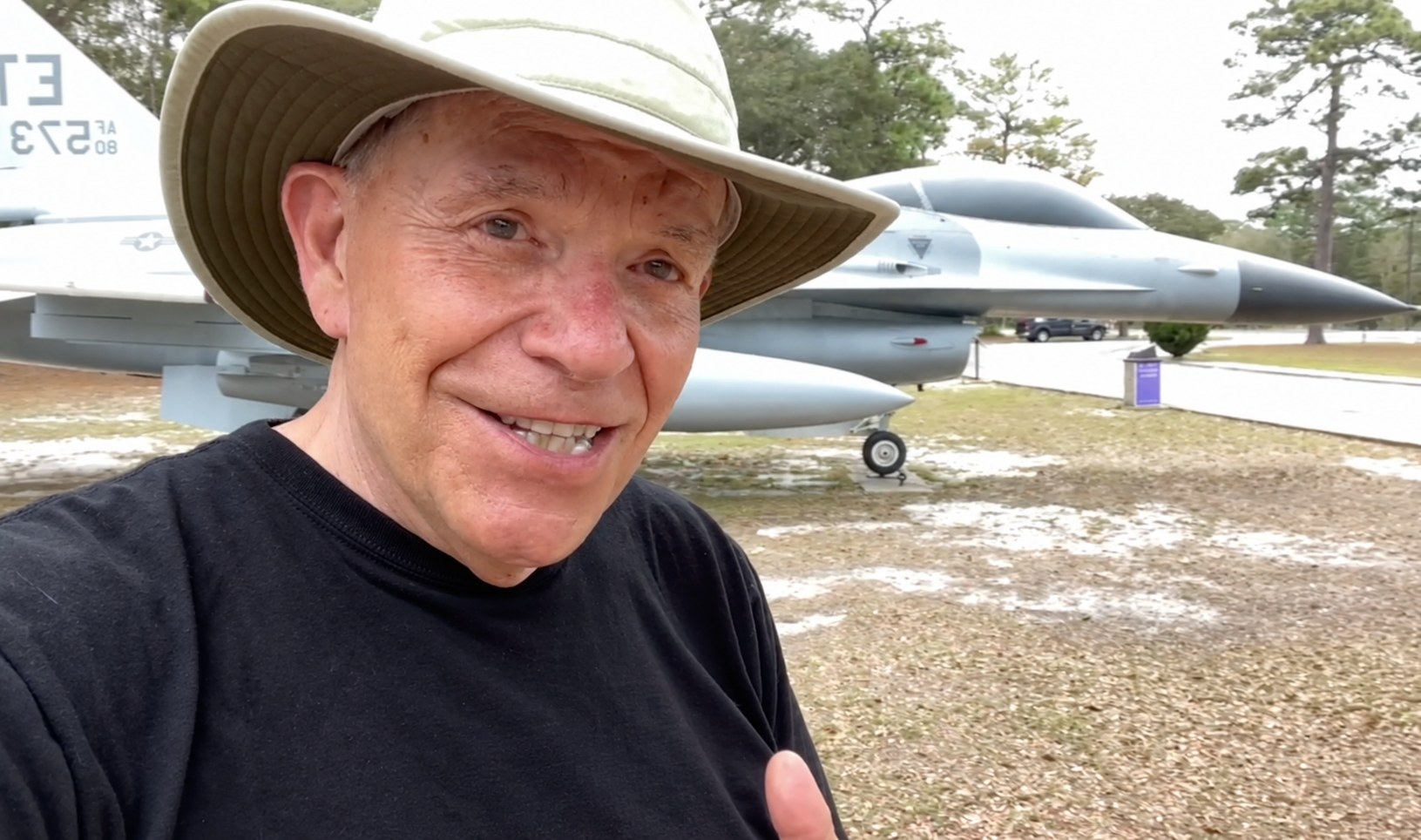 Mike Wendland in a wide-brimmed sun hat, giving a thumbs-up in front of a parked U.S. Air Force jet